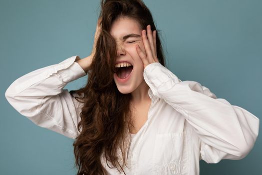 Attractive emotional cute nice adorable tender young curly brunette woman wearing white shirt isolated on blue background with copy space and shouting.