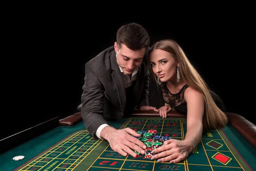 Young beautiful couple takes their winnings at the roulette table at the casino, on a black background. A man in a suit with a woman in a black dress