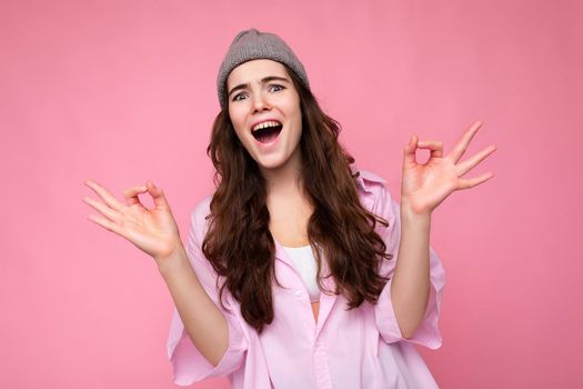 Portrait of young positive happy emotional attractive curly brunette woman with sincere emotions wearing stylish pink shirt and grey hat isolated on pink background with copy space and showing okay gesture and shouting.