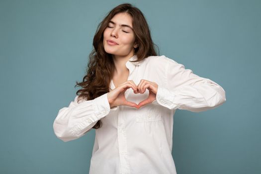 Portrait of young positive happy attractive curly brunette woman with sincere emotions wearing casual white shirt isolated on blue background with free space and making heart form with hands. Love concept.