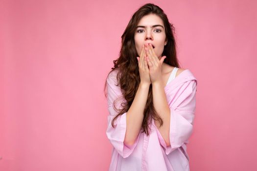 Photo of young shocked amazed surprised winsome nice curly brunette woman with sincere emotions wearing casual pink shirt isolated over pink background with copy space and covering mouth with hands.