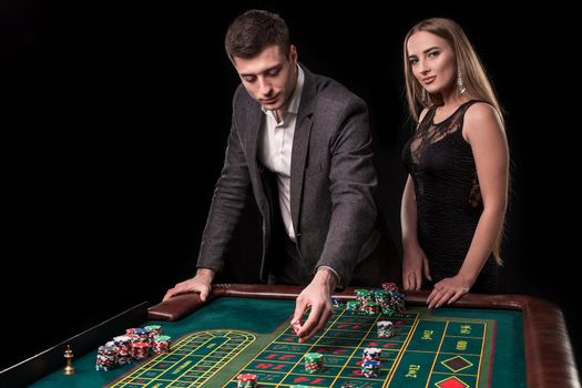 Elegant couple at the casino betting on the roulette, on a black background. A man in a suit with a beautiful young woman in a black dress