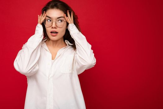 Beautiful young happy surprised curly brunette woman wearing white shirt and optical glasses isolated on red background with copy space. Fashion concept.