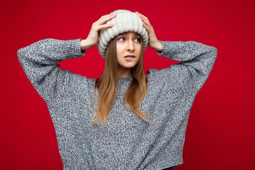 Photo of attractive upset young dark blonde female person standing isolated over red background wall wearing grey sweater and winter beige hat looking to the side and being concerned.