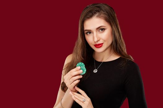 Pretty long hair woman in black dress holding chips for gambling in casino. Studio shot on red background. Poker. Two green Chips in Hand