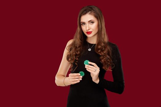 Pretty long hair woman in black dress holding chips for gambling in casino. Studio shot on red background. Poker. Two green Chips in Hand