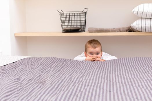 High angle of funny little boy peeking out over opposite side of bed and covering mouth with hands in modern bedroom