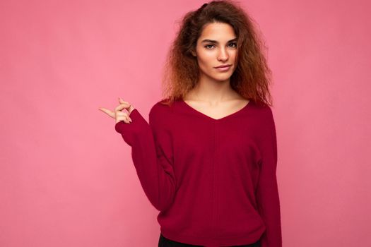 Photo of young beautiful brunette wavy woman with sincere emotions wearing casual pink jersey isolated over pink background with copy space and pointing at free space for mockup.