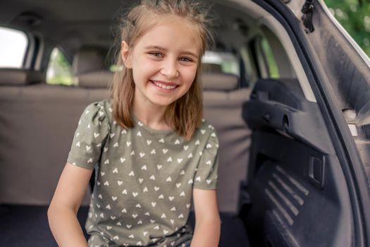 Closeup portrait of cute preteen girl with beautiful hairstyle sitting in the car trunk and smiling looking at the camera. Happy child kid in the vehicle at the nature during summer trip