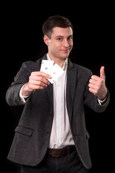 Young caucasian man in a dark suit and a white shirt holding two aces in his hand and showing thumb up hand gesture with smile on black background. Gambling concept. Casino