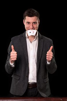 Young caucasian man in a dark suit and a white shirt holding two aces in his teeth and showing thumb up hands gesture on black background. Gambling concept. Casino