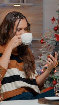 Happy woman using smartphone to talk with family on christmas eve. Young adult talking on video call technology for winter celebration festivity in kitchen with decorations and ornaments