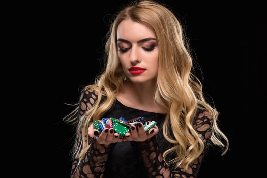 Elegant blonde in a black dress, casino player holding a handful of chips on black background. Poker. Casino. Roulette Blackjack Spin. Caucasian young woman looking at the chips emotions