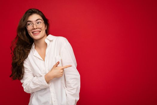 Beautiful happy smiling brunette woman wearing white shirt and optical glasses. Sexy carefree female person posing isolated near red wall in studio with free space. Positive model with natural makeup.