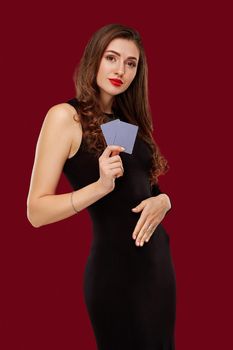 Beautiful confident woman in a sexy black dress showing poker cards, looking at camera with copy place. Studio shot on red background. Casino