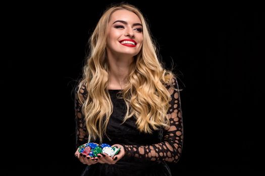Elegant blonde in a black dress, casino player holding a handful of chips on black background. Poker. Casino. Roulette Blackjack Spin. Caucasian young woman looking away emotions