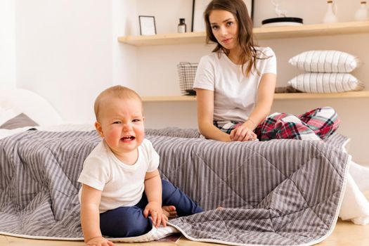 Sad little toddler sitting on floor with young mother in pajamas on bed looking at camera at home