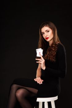 Sexy curly hair brunette in black dress posing with two aces cards in her hands, poker concept on black background. winning combination. sits on a chair