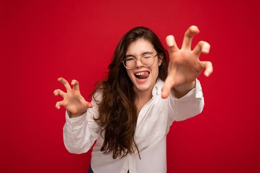 Portrait of young emotional positive sexy beautiful brunette woman with sincere emotions wearing casual white shirt and optical glasses isolated on red background with copy space and making cat claws and growling like animal.