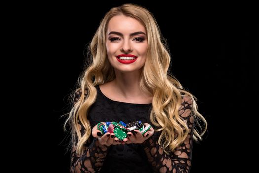 Elegant blonde in a black dress, casino player holding a handful of chips on black background. Poker. Casino. Roulette Blackjack Spin. Caucasian young woman looking at the camera emotions