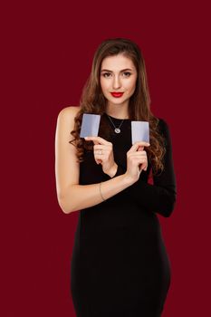 Beautiful confident woman in a sexy black dress showing poker cards, looking at camera with copy place. Studio shot on red background. Casino