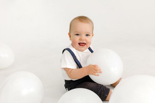 Little vale toddler in white polo and denim jumpsuit laughing while playing with white balloons and looking at camera isolated on white background