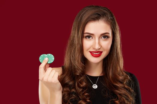 Pretty long hair woman in black dress holding chips for gambling in casino. Studio shot on red background. Poker. Two green Chips in Hand