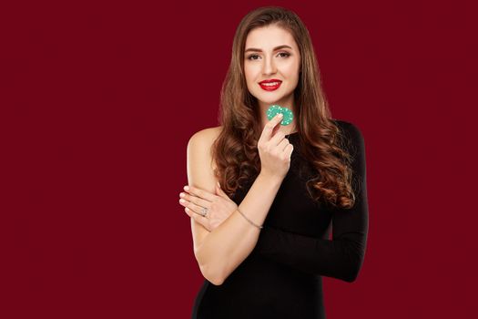 Pretty long hair woman in black dress holding chips for gambling in casino. Studio shot on red background. Poker. Two green Chips in Hand