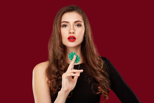 Pretty long hair woman in black dress holding chips for gambling in casino. Studio shot on red background. Poker. Two green Chips in Hand