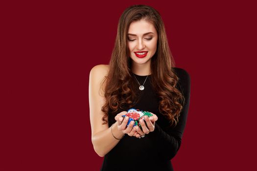 Casino, gambling, poker, people and entertainment concept - woman poker player in black dress with chips in hands on red background. Studio shot