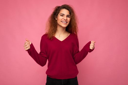 Photo of young happy smiling emotional beautiful brunette curly woman with sincere emotions wearing casual pink pullover isolated over pink background with copy space.