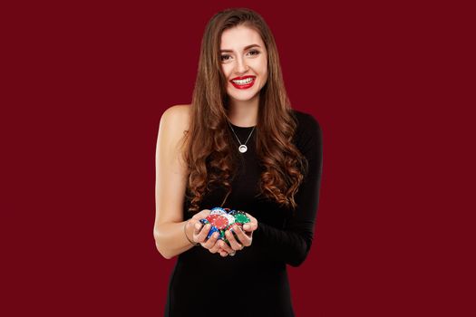 Casino, gambling, poker, people and entertainment concept - woman poker player in black dress with chips in hands on red background. Studio shot