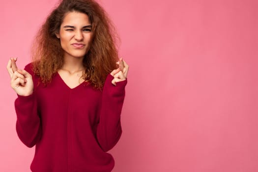 Photo shot of young attractive brunette curly woman with sincere emotions wearing trendy pink pullover isolated over pink background with empty space and keeping crossed fingers. Gesture concept.