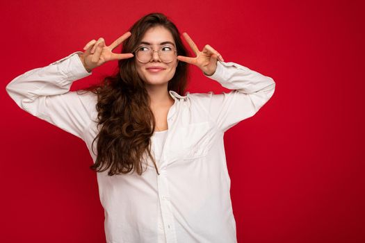 Photo of young beautiful happy smiling brunette woman wearing white shirt and optical glasses. Sexy carefree female person posing isolated near red wall in studio with free space. Positive model with natural makeup.