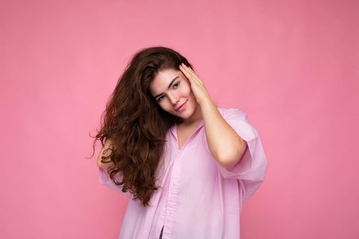 Beautiful charming fascinating young curly brunette woman wearing pink shirt and grey hat isolated on pink background with copy space.