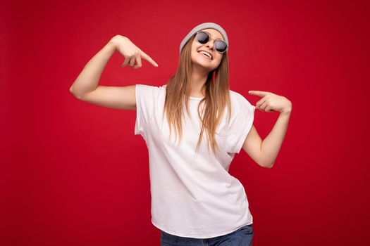 Photo of young amazing positive happy attractive dark blonde woman with sincere emotions wearing casual white t-shirt with empty space for mockup gray hat and sunglasses isolated on red background with copy space and pointing at herself.