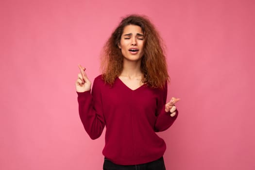 Photo shot of young attractive brunette curly woman with sincere emotions wearing trendy pink sweater isolated over pink background with empty space and holding fingers crossed for good luck. Gesture concept.
