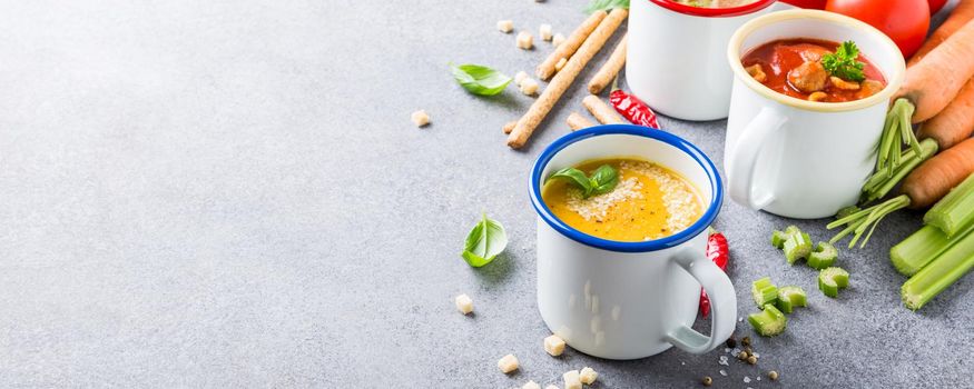 Assorted delicious homemade soups in enamel mugs with ingredients. Healthy food concept with copy space.