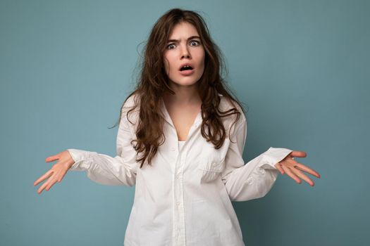 Attractive dissatisfied cute nice adorable tender young curly brunette woman wearing white shirt isolated on blue background with copy space.