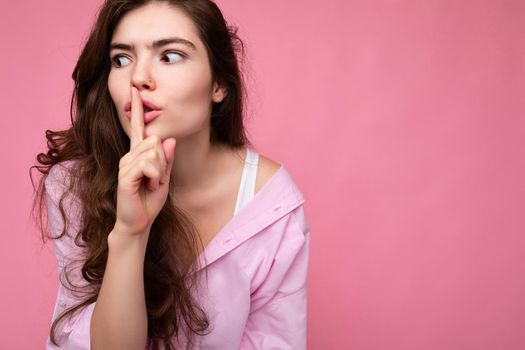 Closeup of young emotional attractive wavy-haired brunette woman with sincere emotions wearing casual pink shirt isolated over pink background with copy space and showing silence gesture with finger near lips. Shhh sign.