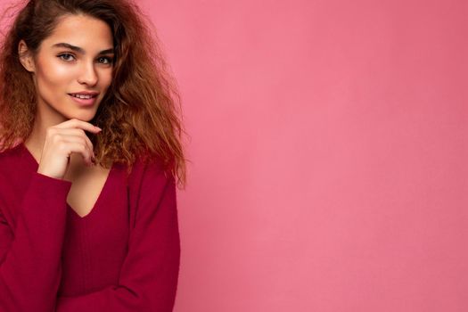 Portrait of young positive thoughtful beautiful brunette curly woman with sincere emotions wearing trendy pink pullover isolated on pink background with copy space.