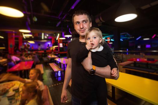 Adult man carrying cute girl and looking away while standing in illuminated cafe of family entertainment center during children party