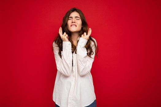 Shot of beautiful emotional young curly brunette woman wearing white shirt isolated on red background with copy space, praying and dreaming.