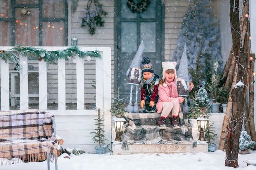 Happy little kids sitting on the porch of the Christmas decorated house, snowing outdoor. Happy New Year and Merry Christmas. Magic winter