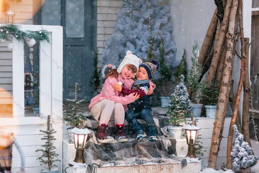 Happy little kids sitting on the porch of the Christmas decorated house, snowing outdoor. Happy New Year and Merry Christmas. Magic winter