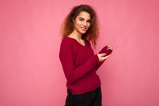 Photo of young happy positive smiling beautiful brunette wavy woman with sincere emotions wearing casual pink jersey isolated over pink background with copy space and pointing at empty space for information.