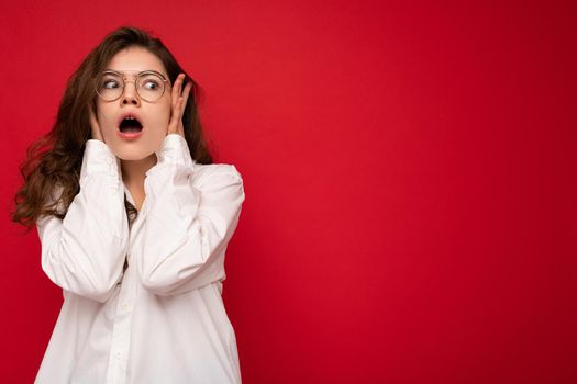 Attractive shocked amazed surprised young curly brunette woman wearing white shirt and optical glasses isolated on red background with empty space.