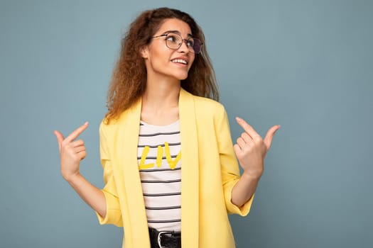 Photo of young positive beautiful brunette curly woman with sincere emotions wearing casual yellow jacket and optical glasses isolated on blue background with copy space and pointing at yourself.