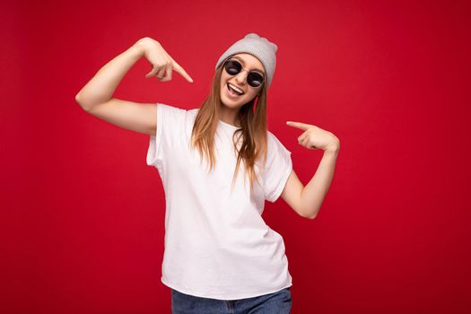 Portrait of young emotional positive happy joyful beautiful dark blonde woman with sincere emotions wearing casual white t-shirt with empty space for mockup gray hat and sunglasses isolated over red background with free space and pointing at herself.