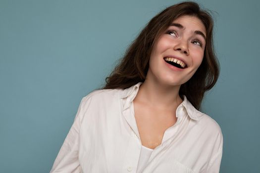 Portrait of beautiful positive cheerful cute smiling young brunette woman in casual white shirt isolated on blue background with copy space.